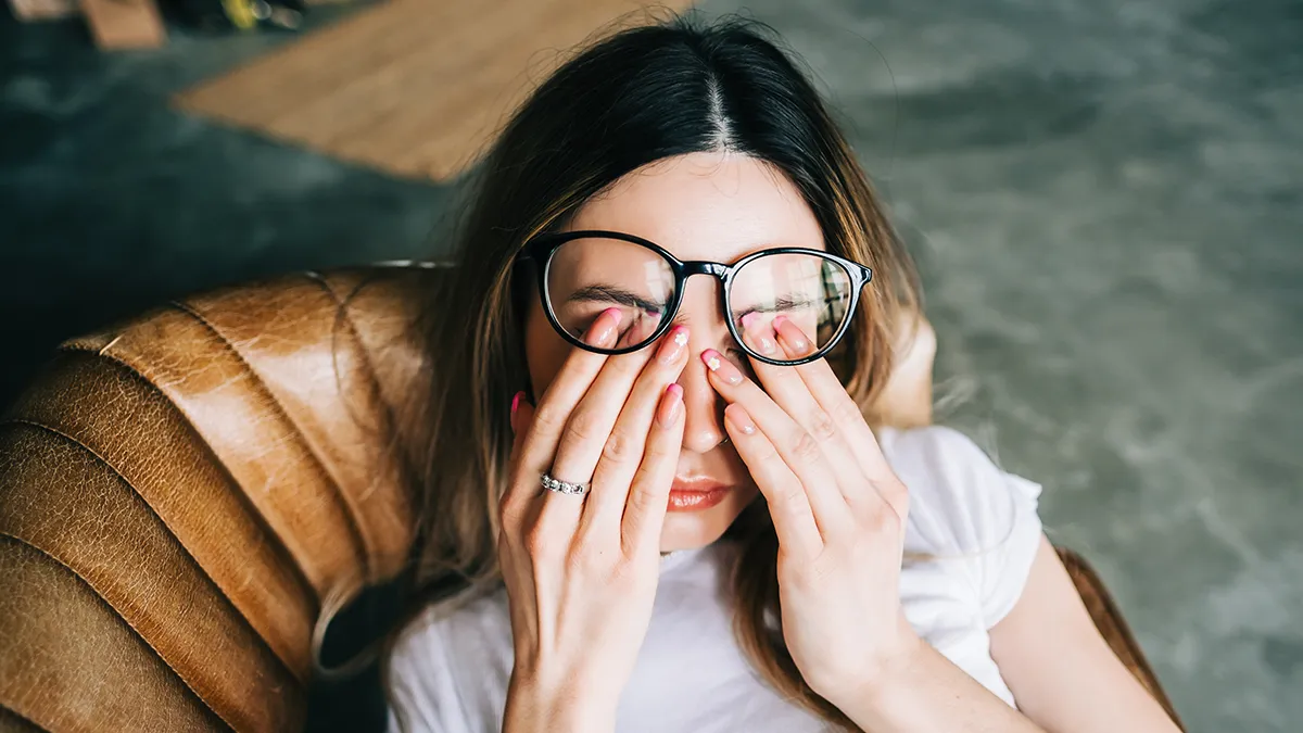 A sleepy woman rubbing her eyes under the glasses in a leather cognac coloured chair